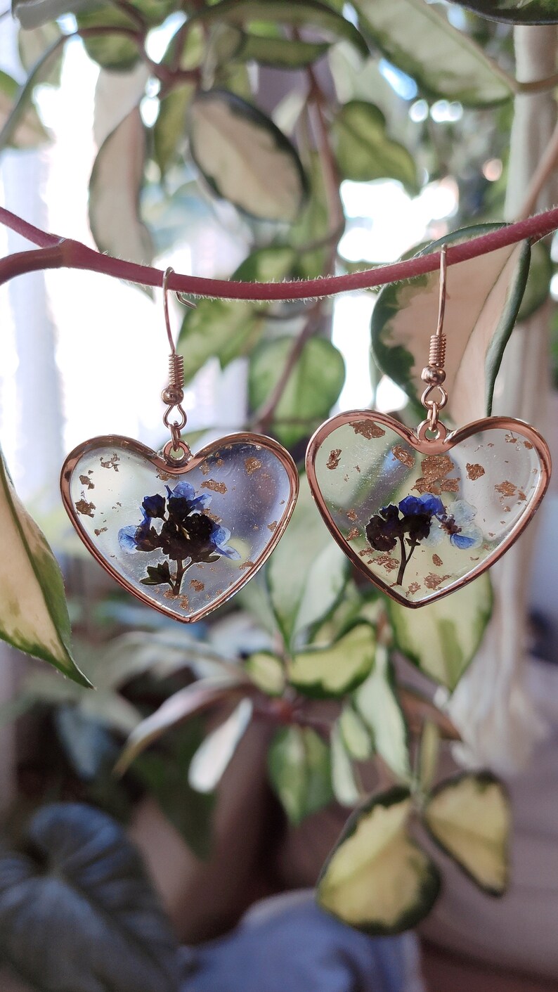 May include: A pair of gold heart-shaped earrings with blue and white pressed flowers and gold flakes inside the resin.