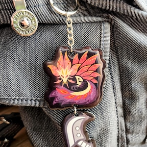 May include: A keychain with a colorful, stylized phoenix design in shades of red, orange, and purple. The keychain also features a smaller charm with a white and gray design, attached by a silver chain and ring. The background is a denim fabric.