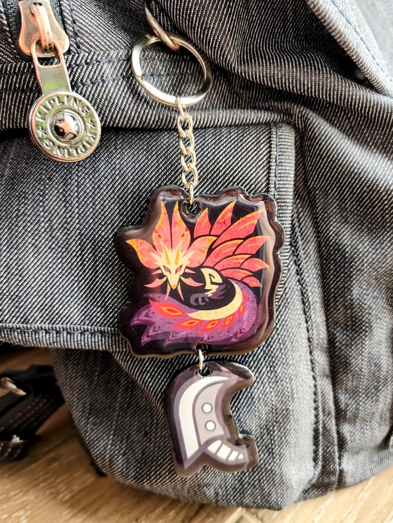 May include: A keychain with a colorful, stylized phoenix design in shades of red, orange, and purple. The keychain also features a smaller charm with a white and gray design, attached by a silver chain and ring. The background is a denim fabric.
