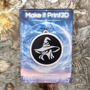 May include: A black and white witch hat keychain. The keychain is round with a black background and a white witch hat design. The card behind the keychain says "Make it Print3D" and "Handmade Keychain".