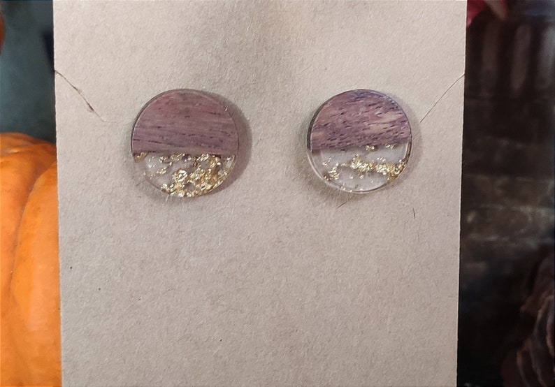 May include: A pair of round wood and resin earrings with gold flakes. The earrings are a light brown colour with a clear resin top half and gold flakes embedded in the resin.
