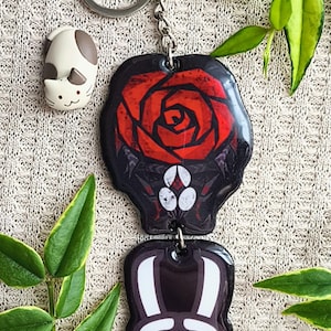 May include: A keychain with two black and red pendants. The top pendant features a red rose design. The bottom pendant has a white and black design. A small cat figurine is also present. The keychain is attached to a silver ring.