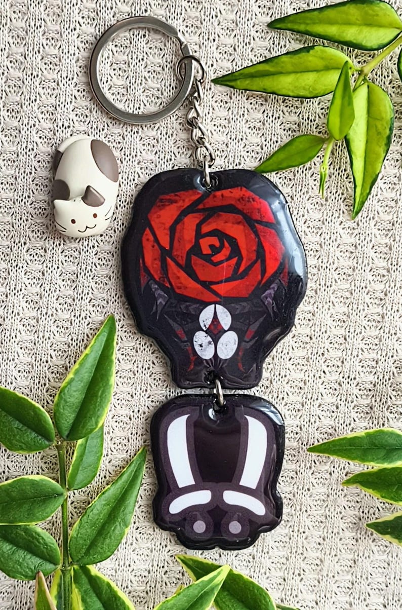 May include: A keychain with two black and red pendants. The top pendant features a red rose design. The bottom pendant has a white and black design. A small cat figurine is also present. The keychain is attached to a silver ring.
