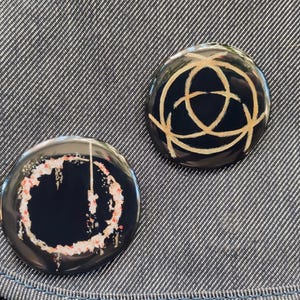 May include: Two round black button pins with gold designs. One pin has a circle with a gold line design. The other pin has a circle with a gold line design and a pink and white splatter pattern.