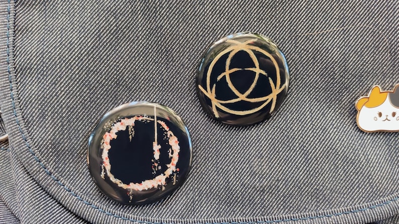 May include: Two round black button pins with gold designs. One pin has a circle with a gold line design. The other pin has a circle with a gold line design and a pink and white splatter pattern.