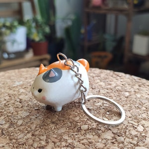 K&ouml;nnte beinhalten: Ein wei&szlig;er, orangefarbener und grauer Keramik-Schl&uuml;sselanh&auml;nger in Form einer Katze mit einer silbernen Kette und einem Ring. Die Katze sitzt auf einer Korkoberfl&auml;che.