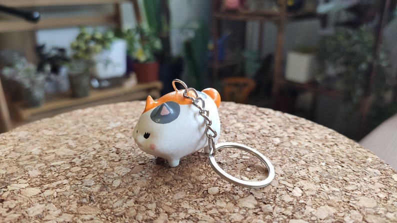 K&ouml;nnte beinhalten: Ein wei&szlig;er, orangefarbener und grauer Keramik-Schl&uuml;sselanh&auml;nger in Form einer Katze mit einer silbernen Kette und einem Ring. Die Katze sitzt auf einer Korkoberfl&auml;che.