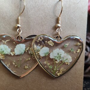 May include: A pair of heart-shaped resin earrings with gold leaf and white pressed flowers. The earrings have gold hooks.