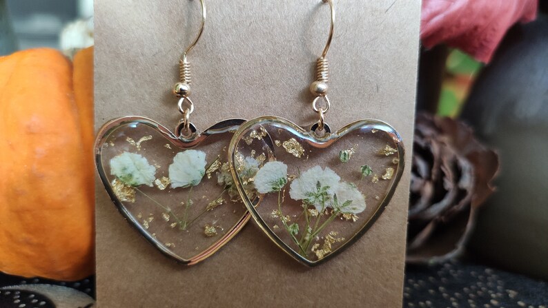 May include: A pair of heart-shaped resin earrings with gold leaf and white pressed flowers. The earrings have gold hooks.