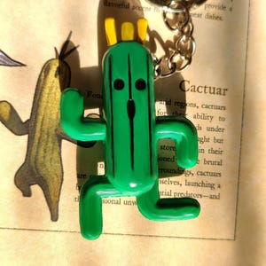 May include: Green Cactuar keychain with a yellow crown and black eyes. The keychain is made of plastic and is a popular character from the Final Fantasy video game series.