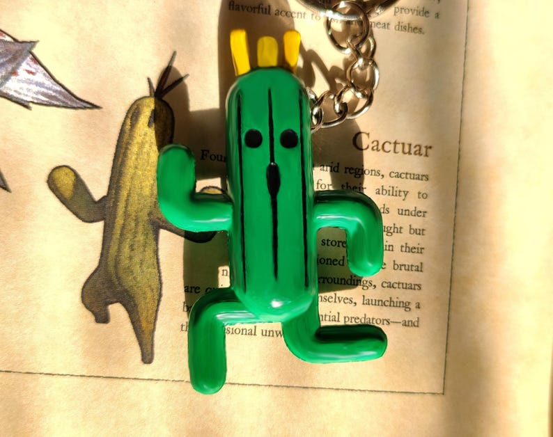 May include: Green Cactuar keychain with a yellow crown and black eyes. The keychain is made of plastic and is a popular character from the Final Fantasy video game series.