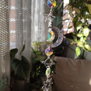 May include: A celestial-themed suncatcher with a silver chain. It features a star, crescent moon, planet, and sun charms, along with iridescent crystal accents. The suncatcher is designed to hang and catch the light.