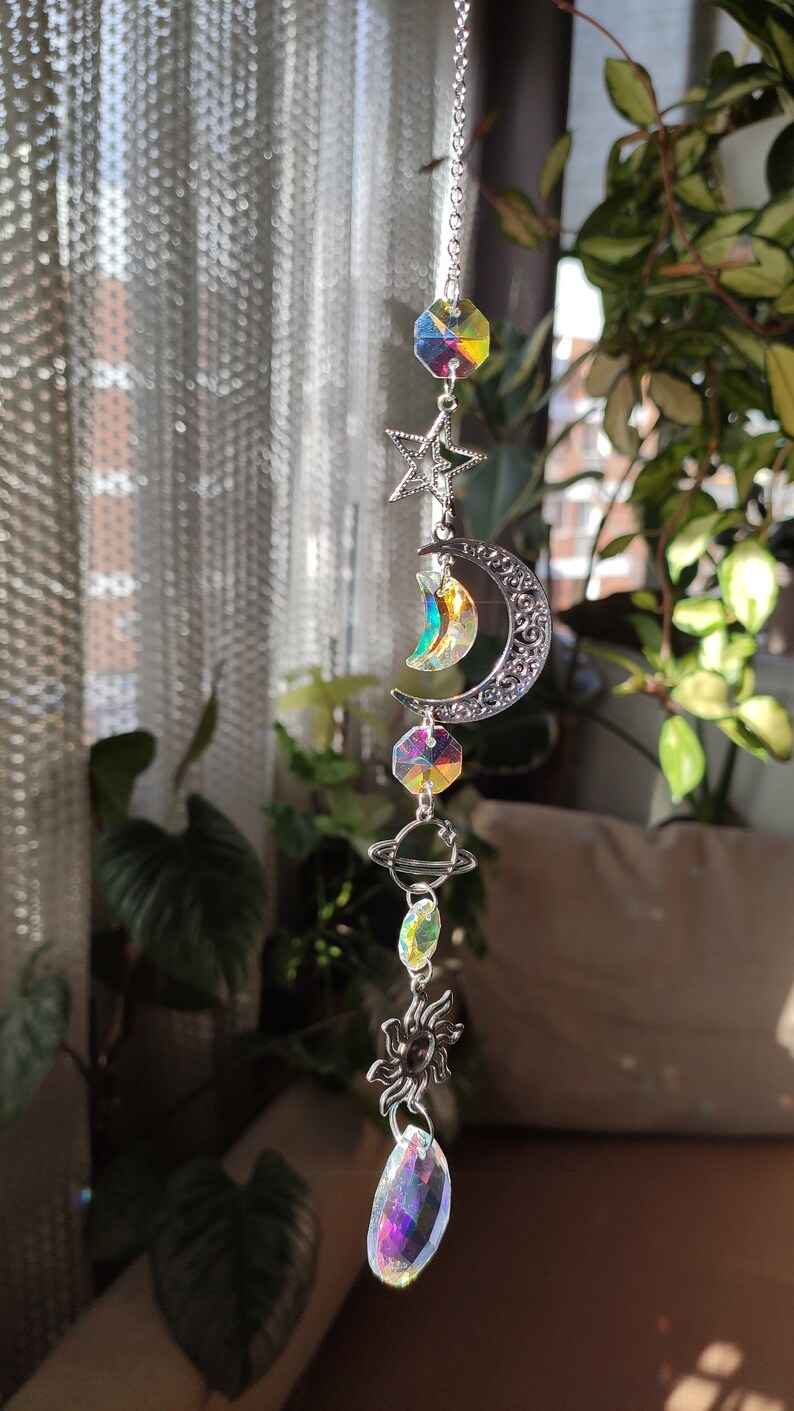 May include: A celestial-themed suncatcher with a silver chain. It features a star, crescent moon, planet, and sun charms, along with iridescent crystal accents. The suncatcher is designed to hang and catch the light.