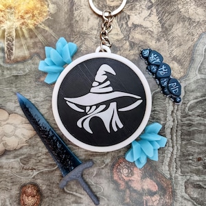 May include: A black and white keychain with a wizard hat design, a blue sword, and blue floral accents. The keychain is attached to a silver keyring. The background features a map design.