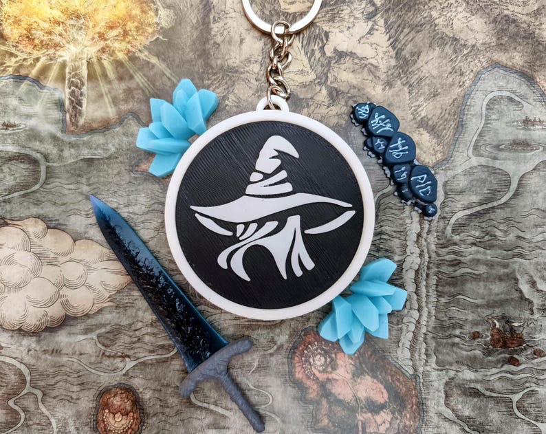 May include: A black and white keychain with a wizard hat design, a blue sword, and blue floral accents. The keychain is attached to a silver keyring. The background features a map design.