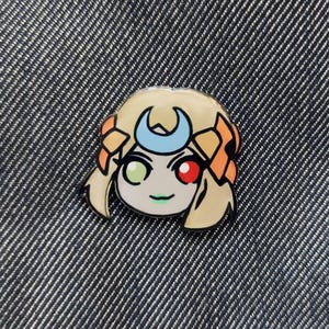 May include: A yellow and orange enamel pin featuring a cartoon character with a crescent moon headband, green and red eyes, and a smile.