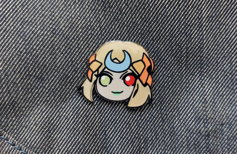 May include: A yellow and orange enamel pin featuring a cartoon character with a crescent moon headband, green and red eyes, and a smile.