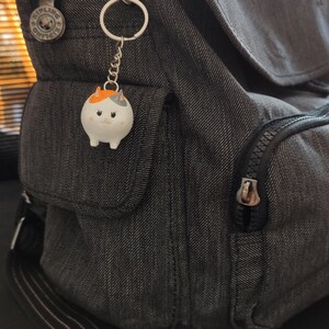 K&ouml;nnte beinhalten: Ein grauer Rucksack mit einer Rei&szlig;verschlusstasche und einem kleinen wei&szlig;en Katzen-Schl&uuml;sselanh&auml;nger mit orangefarbenen Ohren und einer schwarzen Nase.