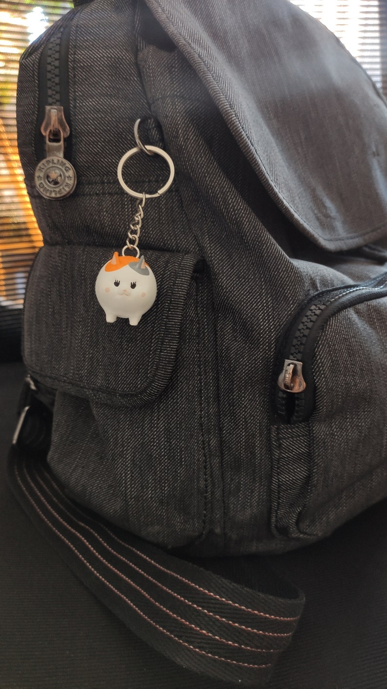 K&ouml;nnte beinhalten: Ein grauer Rucksack mit einer Rei&szlig;verschlusstasche und einem kleinen wei&szlig;en Katzen-Schl&uuml;sselanh&auml;nger mit orangefarbenen Ohren und einer schwarzen Nase.