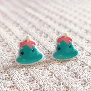 May include: A pair of whimsical stud earrings. Each earring features a teal, cartoon-like character with a red and yellow top. The earrings are set against a white, textured background.
