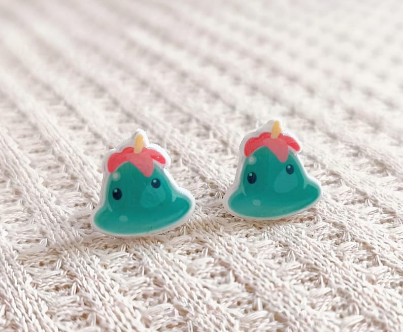 May include: A pair of whimsical stud earrings. Each earring features a teal, cartoon-like character with a red and yellow top. The earrings are set against a white, textured background.