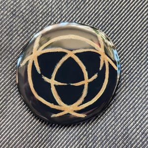 May include: A round, black button with a gold-colored design of three overlapping circles. The button is on a gray fabric background.