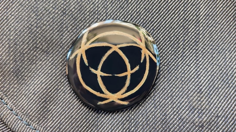 May include: A round, black button with a gold-colored design of three overlapping circles. The button is on a gray fabric background.