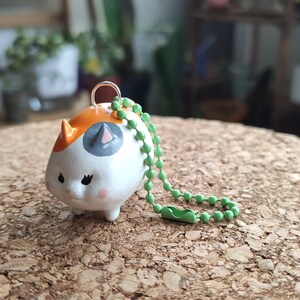 K&ouml;nnte beinhalten: Ein kleiner Keramik-Schl&uuml;sselanh&auml;nger in Form einer Katze mit orangefarbenen, wei&szlig;en und grauen Markierungen. Der Schl&uuml;sselanh&auml;nger hat eine gr&uuml;ne Perlenkette.
