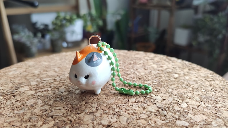 K&ouml;nnte beinhalten: Ein kleiner Keramik-Schl&uuml;sselanh&auml;nger in Form einer Katze mit orangefarbenen, wei&szlig;en und grauen Markierungen. Der Schl&uuml;sselanh&auml;nger hat eine gr&uuml;ne Perlenkette.