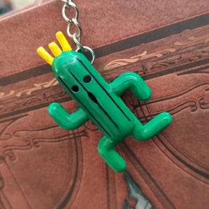 May include: A green cactus character keychain with a yellow crown. The character has a black face with two eyes and a mouth. The keychain is attached to a silver chain.