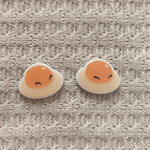 May include: Two cute, smiling fried egg patches with orange yolks and white egg whites. The patches are on a textured, light beige fabric background. These patches are perfect for adding to clothing or accessories.