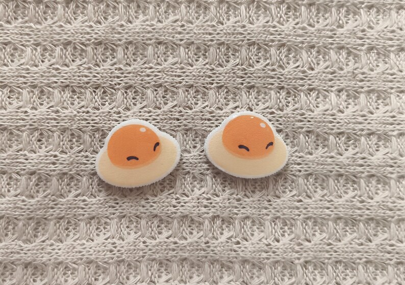 May include: Two cute, smiling fried egg patches with orange yolks and white egg whites. The patches are on a textured, light beige fabric background. These patches are perfect for adding to clothing or accessories.
