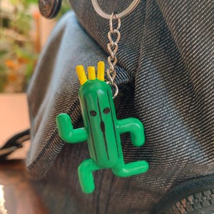 May include: A green cactus character keychain with a yellow crown, black eyes, and a black line down the center. The keychain is attached to a silver chain and a silver ring.