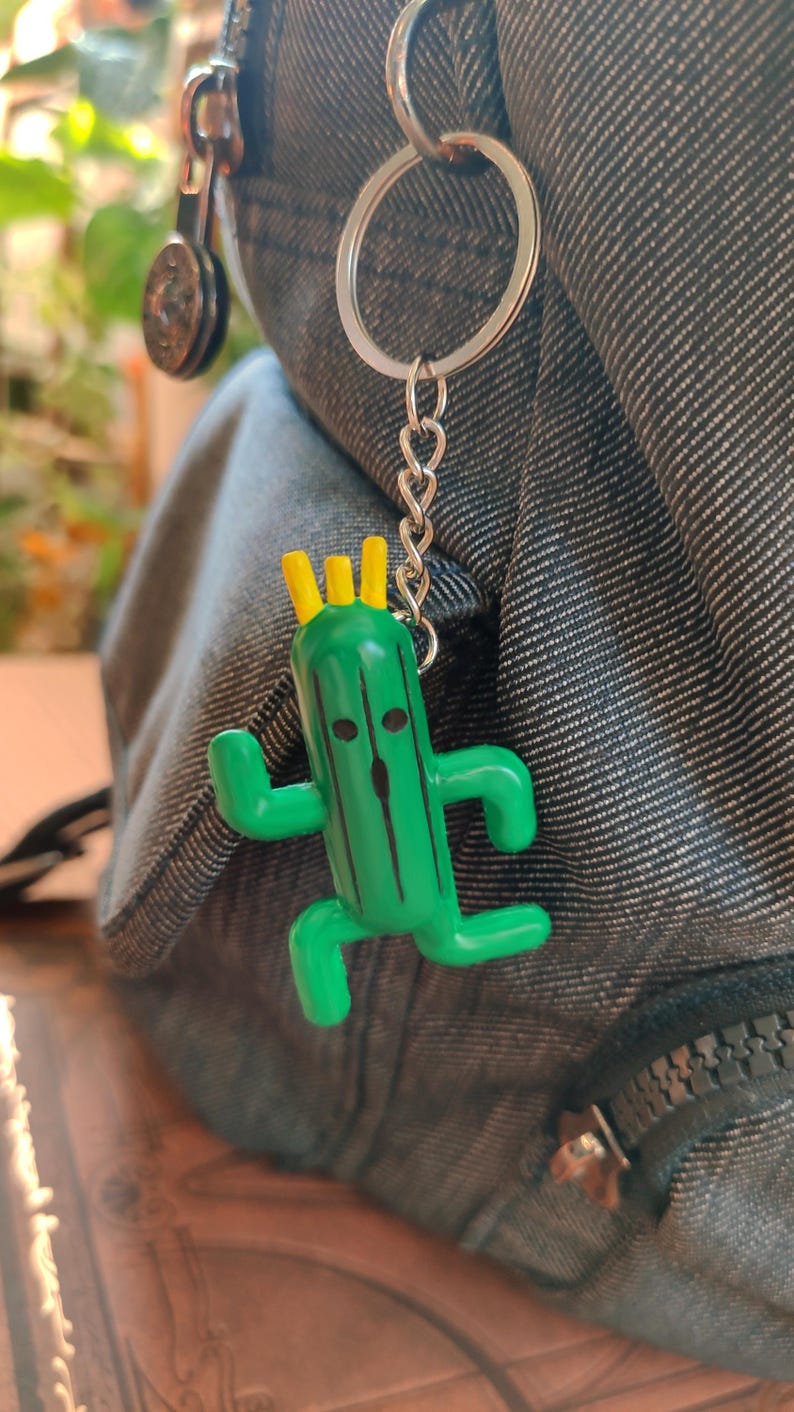 May include: A green cactus character keychain with a yellow crown, black eyes, and a black line down the center. The keychain is attached to a silver chain and a silver ring.