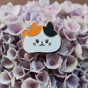 May include: A white enamel pin with a cartoon cat design. The cat has orange, black, and white fur and a cute expression. The pin is on a background of purple flowers.