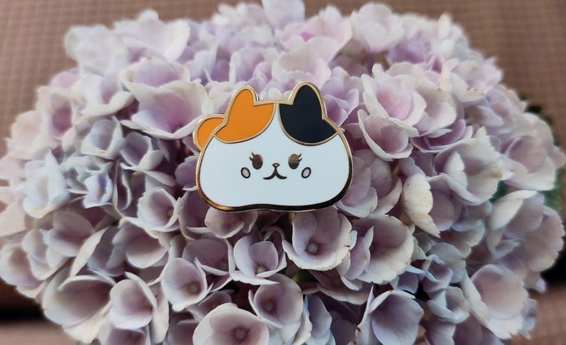May include: A white enamel pin with a cartoon cat design. The cat has orange, black, and white fur and a cute expression. The pin is on a background of purple flowers.