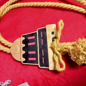 May include: A gold rope necklace with a black and red pendant shaped like a building. The pendant has a gold trim and a small gold circle at the top.