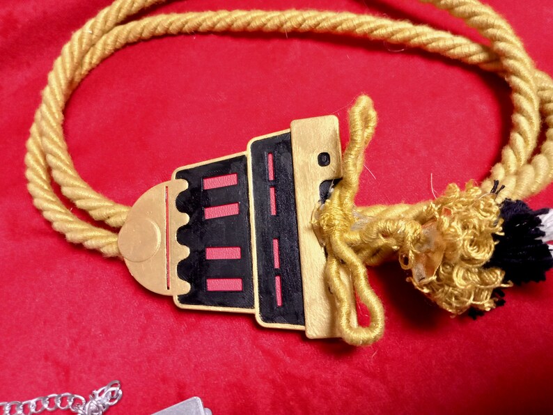 May include: A gold rope necklace with a black and red pendant shaped like a building. The pendant has a gold trim and a small gold circle at the top.
