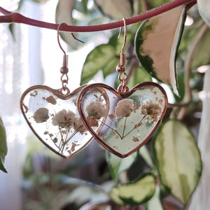 May include: A pair of gold heart-shaped earrings with delicate white flowers pressed inside the resin. The earrings have a gold hook closure.