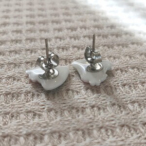 May include: A pair of white ceramic stud earrings with silver-colored posts and butterfly backs. The earrings are in the shape of a stylized bird. The earrings are displayed on a textured, light-colored surface.