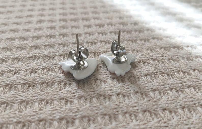 May include: A pair of white ceramic stud earrings with silver-colored posts and butterfly backs. The earrings are in the shape of a stylized bird. The earrings are displayed on a textured, light-colored surface.