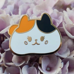May include: A white enamel pin with a cartoon cat design. The cat has orange and black fur and a cute expression. The pin is surrounded by purple flowers.