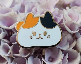 Puede incluir: Un pin de esmalte blanco con un diseño de gato de dibujos animados. El gato tiene un pelaje naranja y negro y una expresión linda. El pin está rodeado de flores moradas.