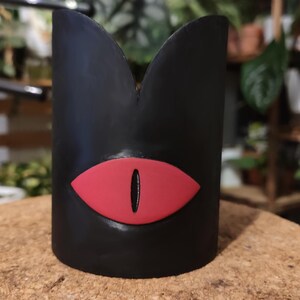 Op de afbeelding: Zwarte keramische plantenpot met een rood oogontwerp. De pot heeft de vorm van een hoge cilinder met een afgeronde bovenkant.