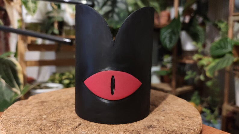 Op de afbeelding: Zwarte keramische plantenpot met een rood oogontwerp. De pot heeft de vorm van een hoge cilinder met een afgeronde bovenkant.