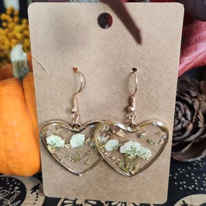 May include: A pair of gold heart-shaped earrings with white pressed flowers and gold flakes inside the resin.