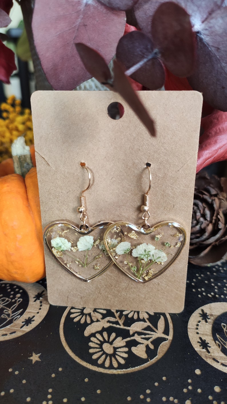 May include: A pair of gold heart-shaped earrings with white pressed flowers and gold flakes inside the resin.