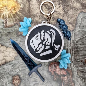 May include: A collection of fantasy-themed accessories, including a keychain with a black and white emblem, a blue sword, and blue floral accents. The items are arranged on a map-like background.