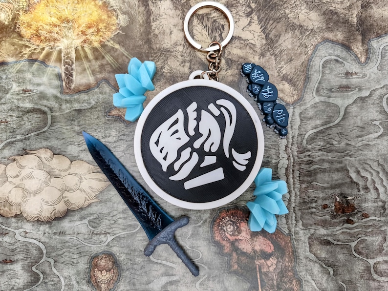 May include: A collection of fantasy-themed accessories, including a keychain with a black and white emblem, a blue sword, and blue floral accents. The items are arranged on a map-like background.
