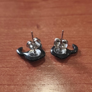 May include: A pair of black stud earrings with a mustache design. The earrings have a silver post and backing. The earrings are on a brown wooden surface.